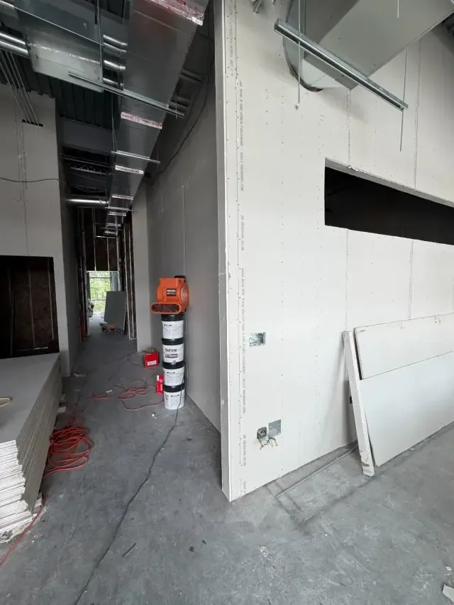 Interior corridor under construction with newly hung drywall, a framed pass-through opening, and overhead ductwork.