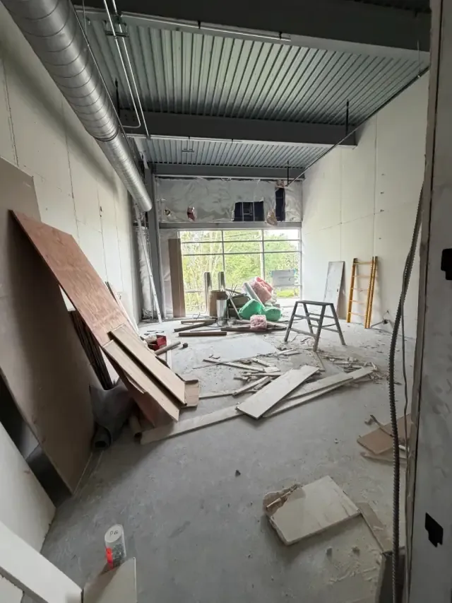 Wide view of a future studio room under construction, with a large storefront window letting in daylight and drywall staged on the floor.
