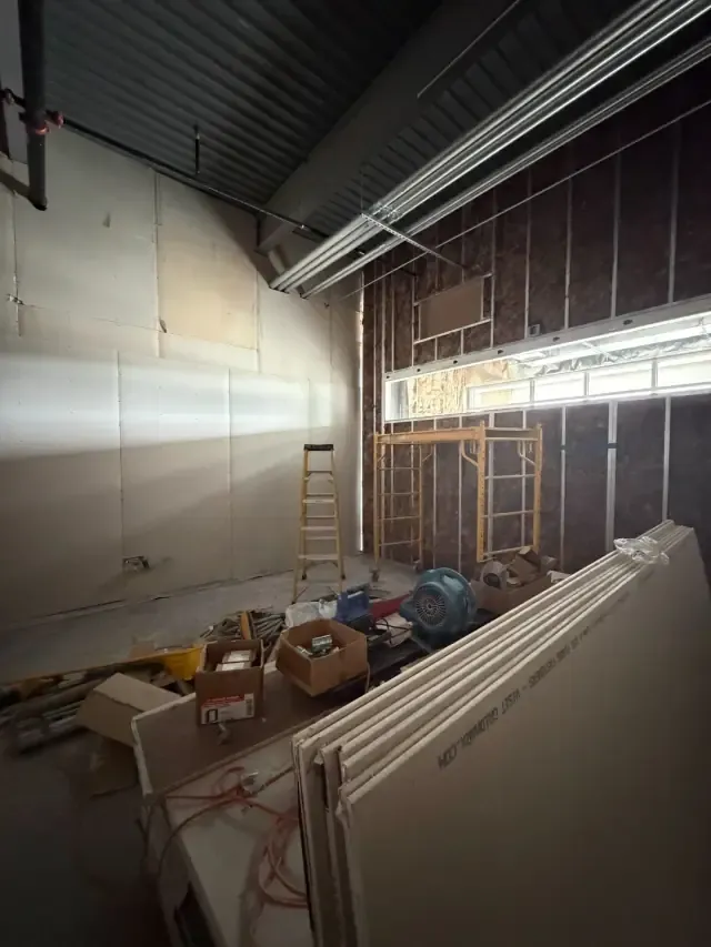 Studio room mid-build with a long transom window, scaffold, ladder, and stacked sound-control drywall.